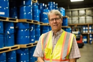 Bill stands in a warehouse of blue barrels wearing a yellow reflective vest.