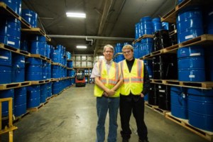 Bill and Cody stand in a warehouse full of blue barrels, both smiling and wearing reflective vests, Bill crosses his hands in front of him and Cody has his hands in his pockets.