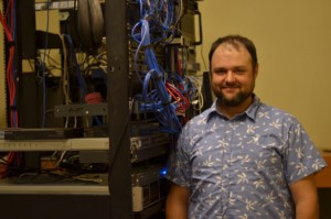 Sean stands next to a wall of wires and hardware, smiling for the camera.