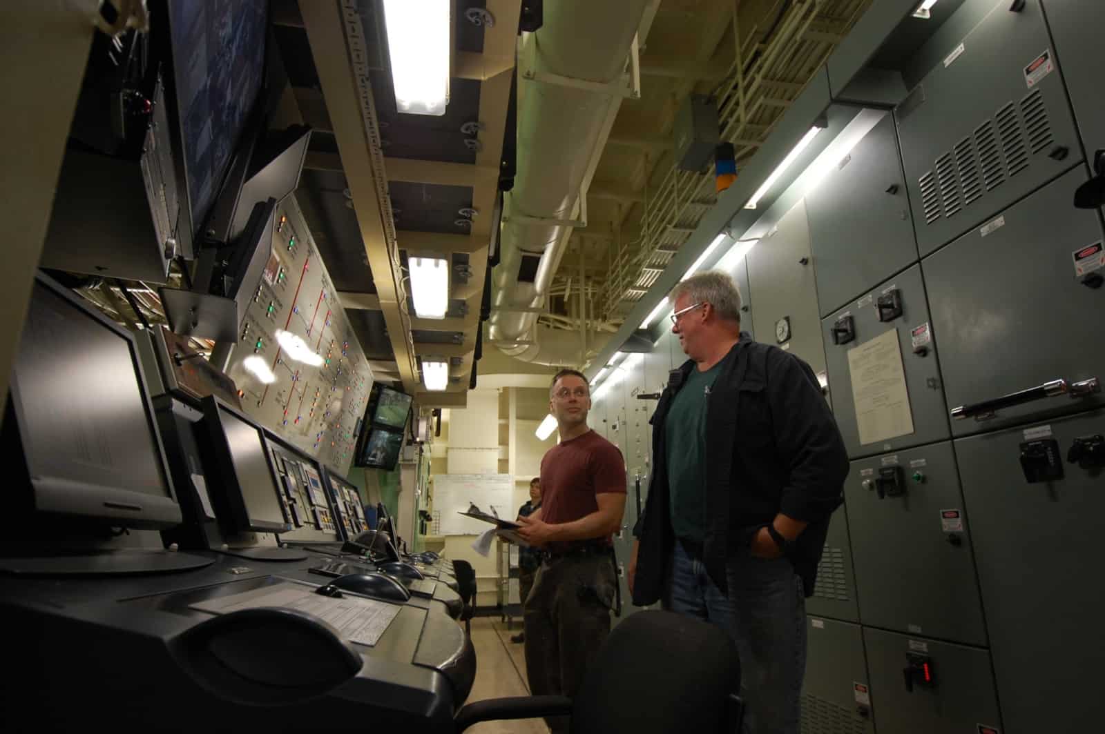 Hary Poole stands in a ship's engine room talking to a worker