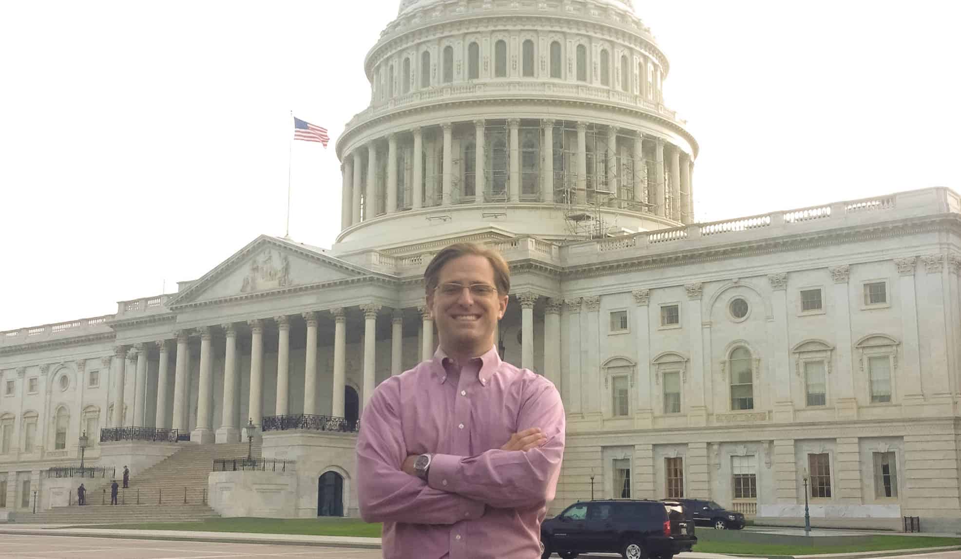 Chris Coakley stands with his arms crossed smiling in front of Washington D.C.'s capitol building.