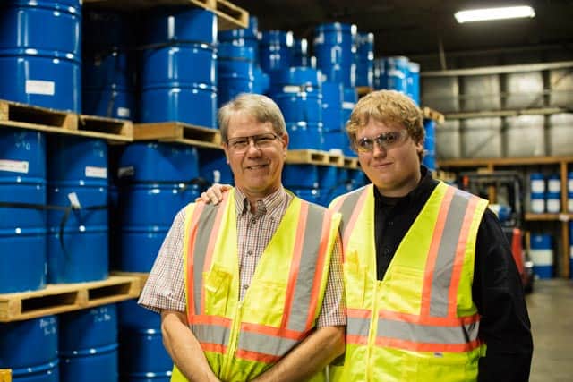 Bill and Cody smile for the camera in a warehouse full of blue barrels, both smile and wear a yellow reflective vest, Cody rests his arm over Bill's shoulder.