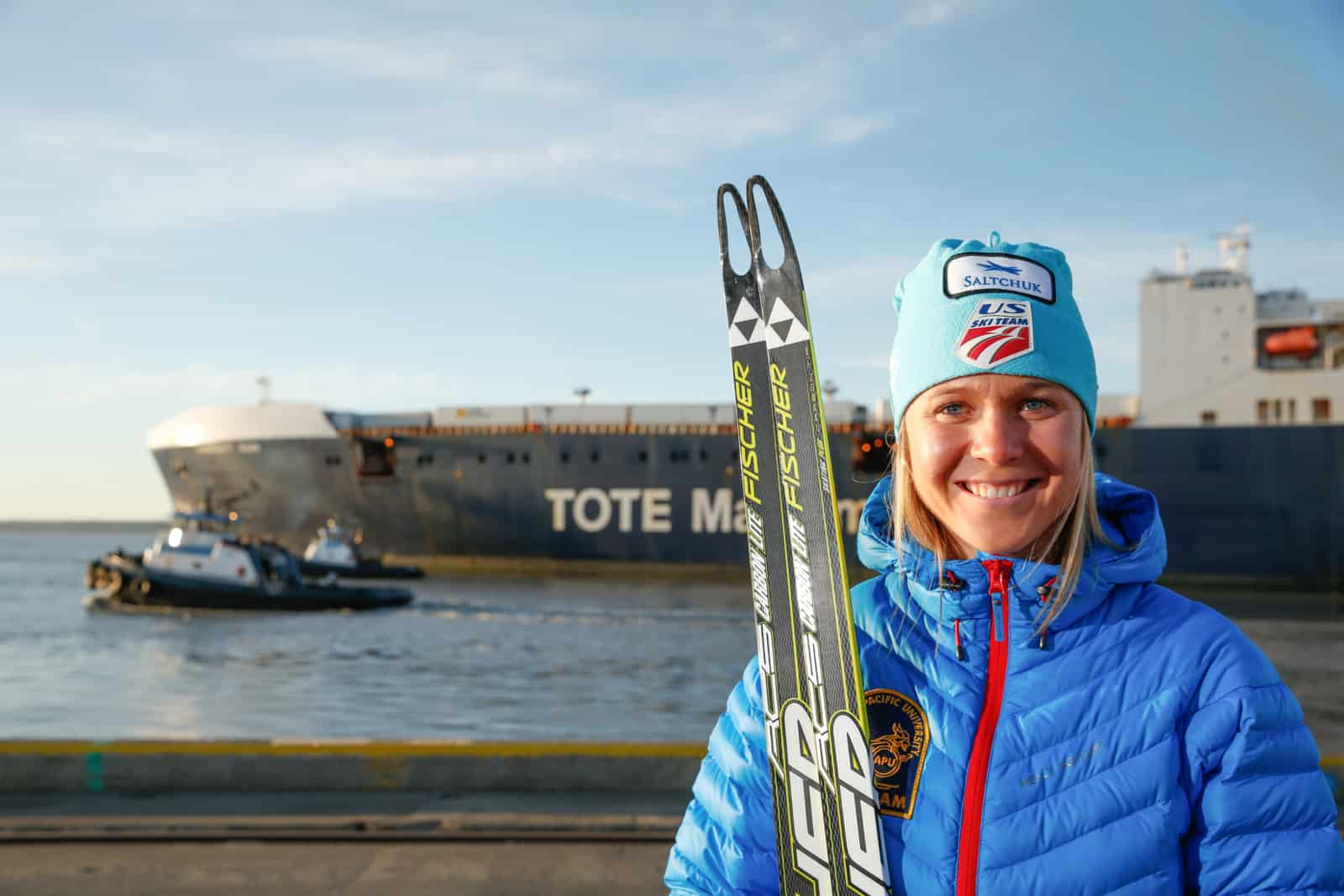 Sadie holds her skis in a puff coat in front of a TOTE maritime ship.