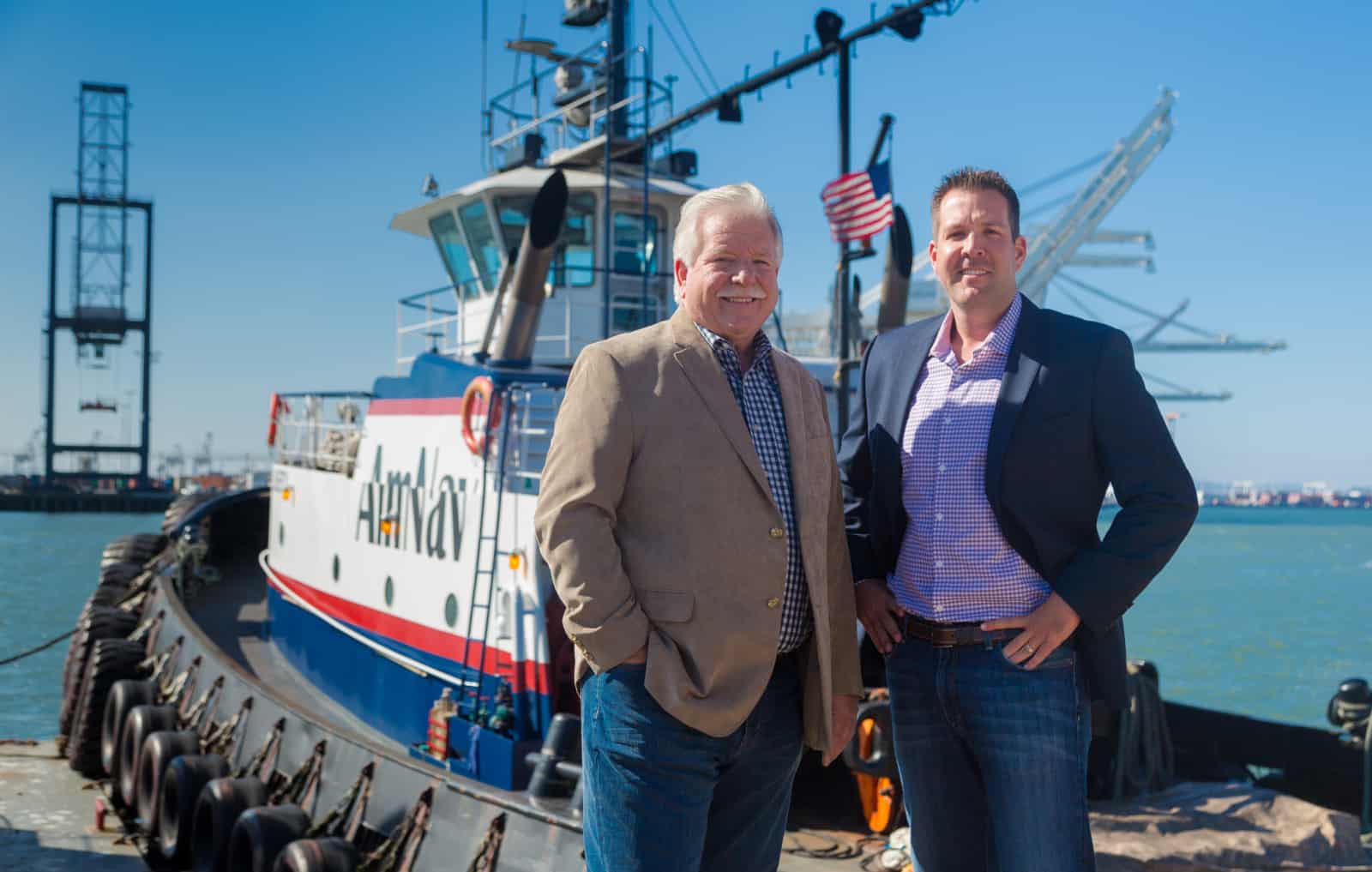 Milt and Dave Merrit stand in front of an AmNav tractor tugboat, both wearing casual suits.