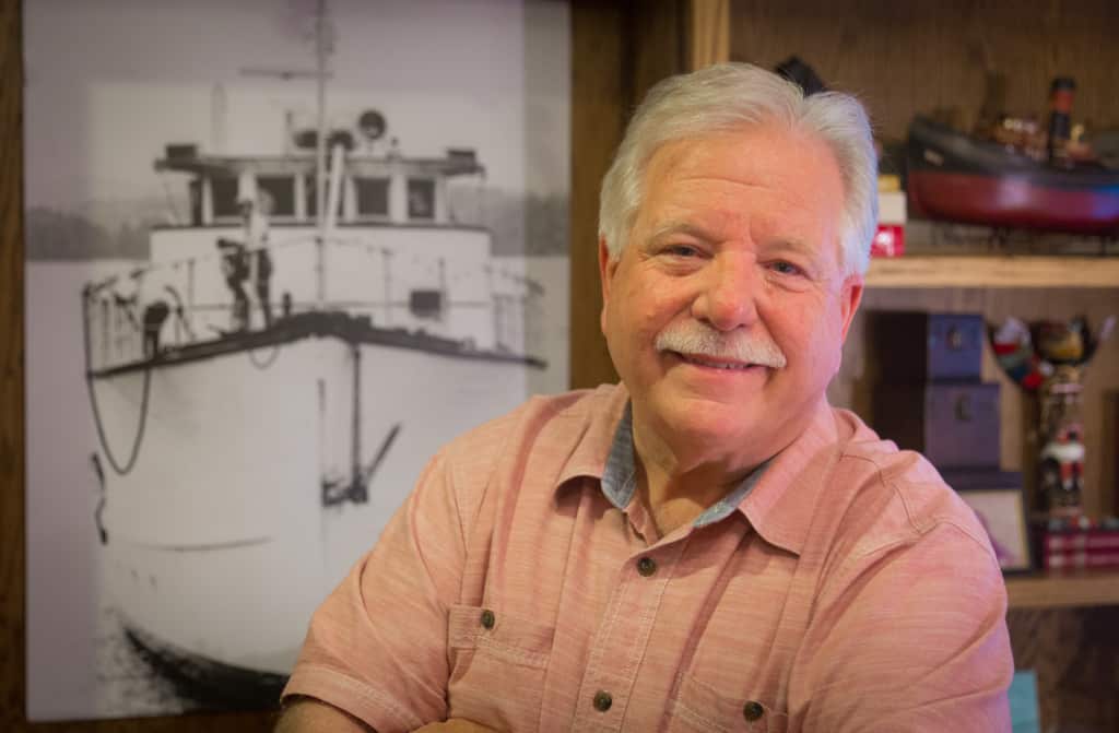 Milt Merritt in his home office, with a framed photo of the M/V Willis Shank behind him.