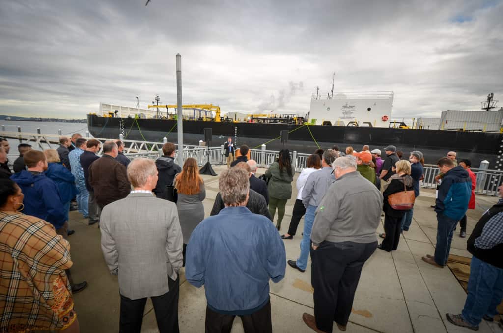 A crowd faces a new barge, listening to a speaker talk about Antril.