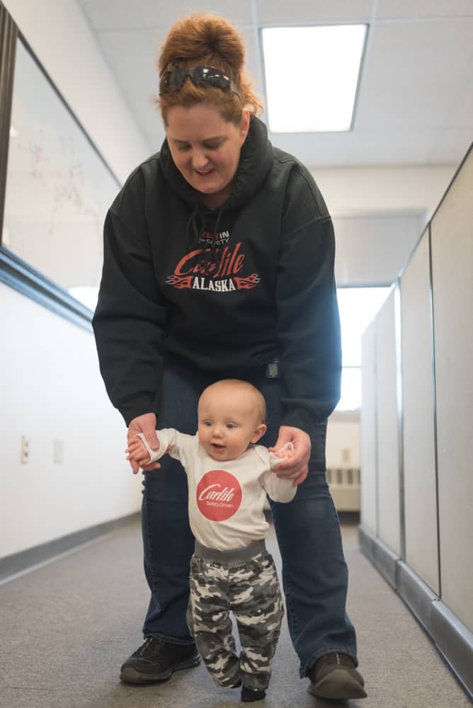 Sprinkle, wearing a Carlile hoodie, holds the hands of her baby grandson, wearing a Carlile shirt, helping him walk in her office.