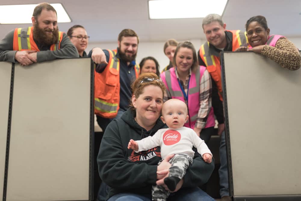 Sprinkle holds her baby grandson and smiles for the camera, a group of Carlile employees gathers around her.