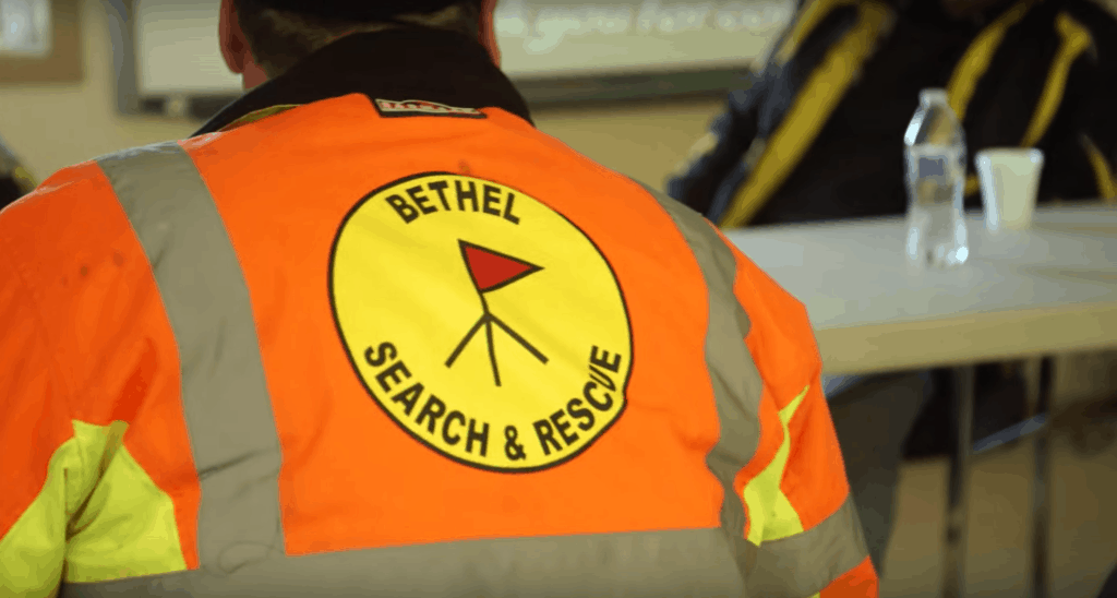 The back of someone wearing an orange and yellow reflective Bethel Search & Rescue jacket.