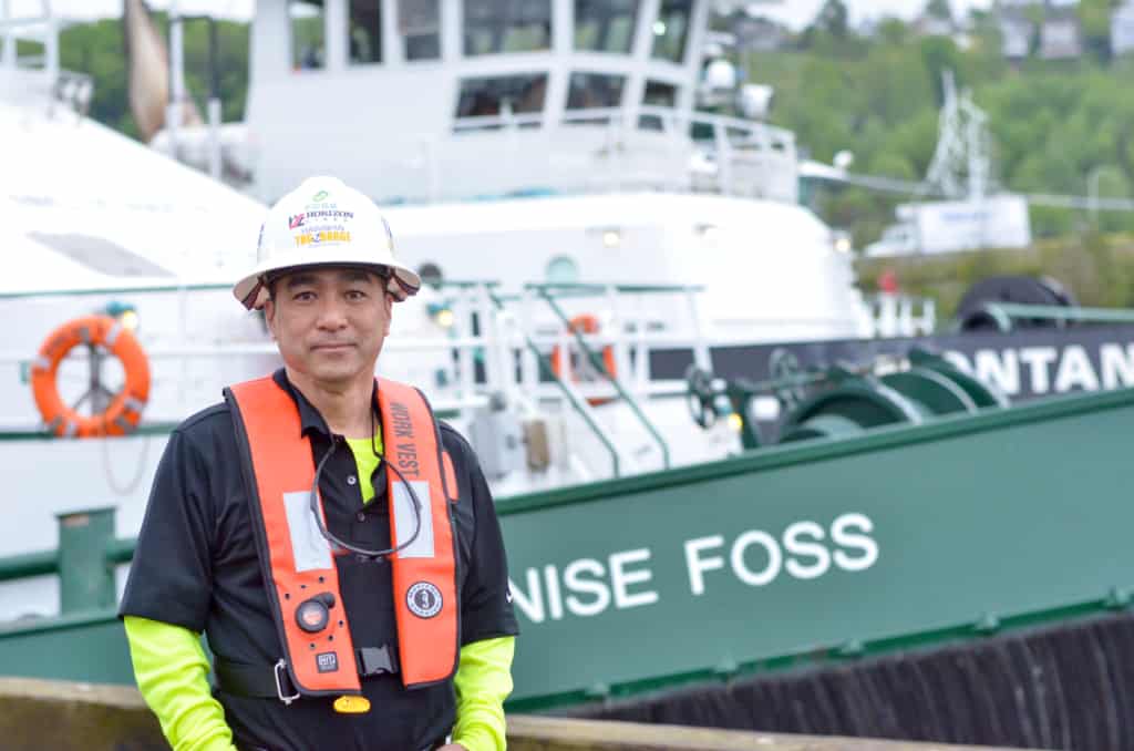 Lau stands in front of a Foss tugboat in safety hard hat, orange life vest, and neon shirt.