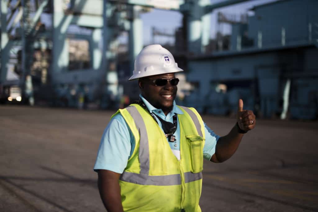 Williams stands in a shipyard smiling and giving a thumbs up, he has on a green reflective vest and TOTE hard hat.