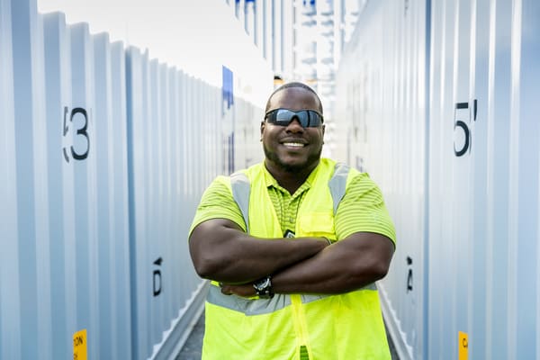 Williams stands with arms crossed and a smile on his face between TOTE shipping containers, Williams is wearing a green reflective vest and wraparound sunglasses.