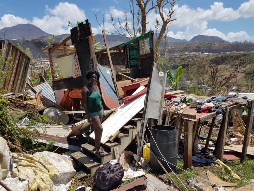 Dominican Tropical employee stands at the base of stairs leading to her broken home. Devastation is seen all around.