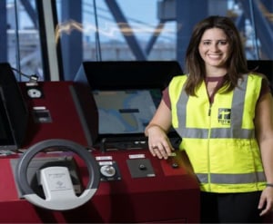 Yahaira leans on a console for some heavy machinery smiling, wearing a TOTE Maritime reflective vest.