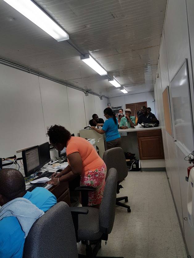 An impromptu welcome desk and workstations are set up in a small space with fluorescent lights, several people wait for help as Tropical employees are hard at work.