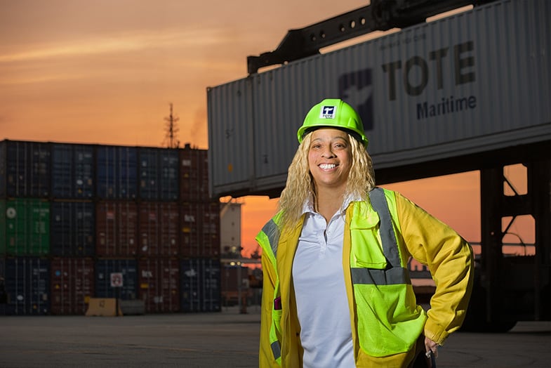 Coleman smiles with hand on hip wearing a green reflective vest and hard hat. The sun sets on a TMPR shipyard behind her.