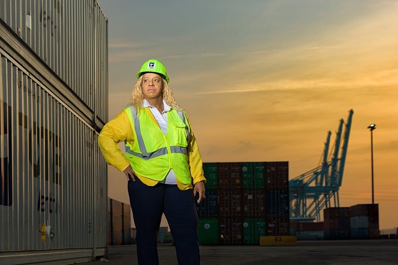 Coleman poses with hand on hip in a green reflective vest and green TOTE hard hat, behind her the sun sets on a TMPR shipyard.