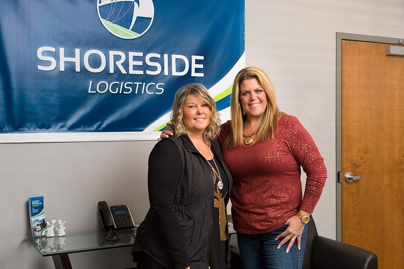 Stephanie and Stacey pose in front of a Shoreside Logistics banner.