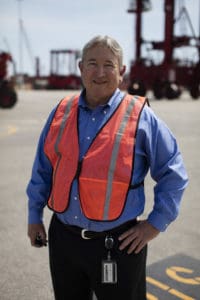 Culpepper, wearing an orange reflective vest, stands with hand on hip in a shipyard.