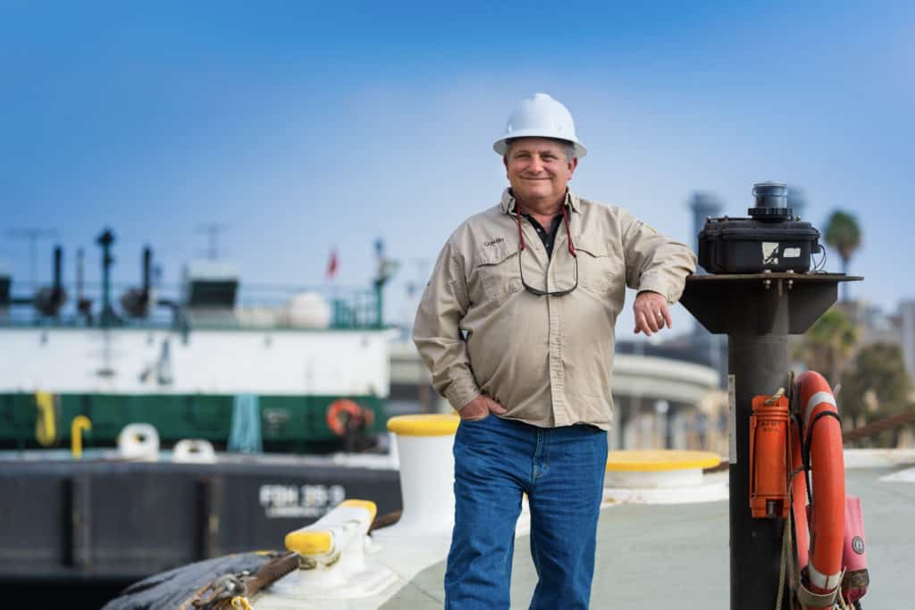 Ron, smiling, leans on a pillar on a barge wearing a hard hat.