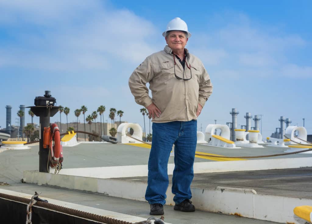 Ron proudly stands on a barge with hand on his hips, wearing a white hard hat.