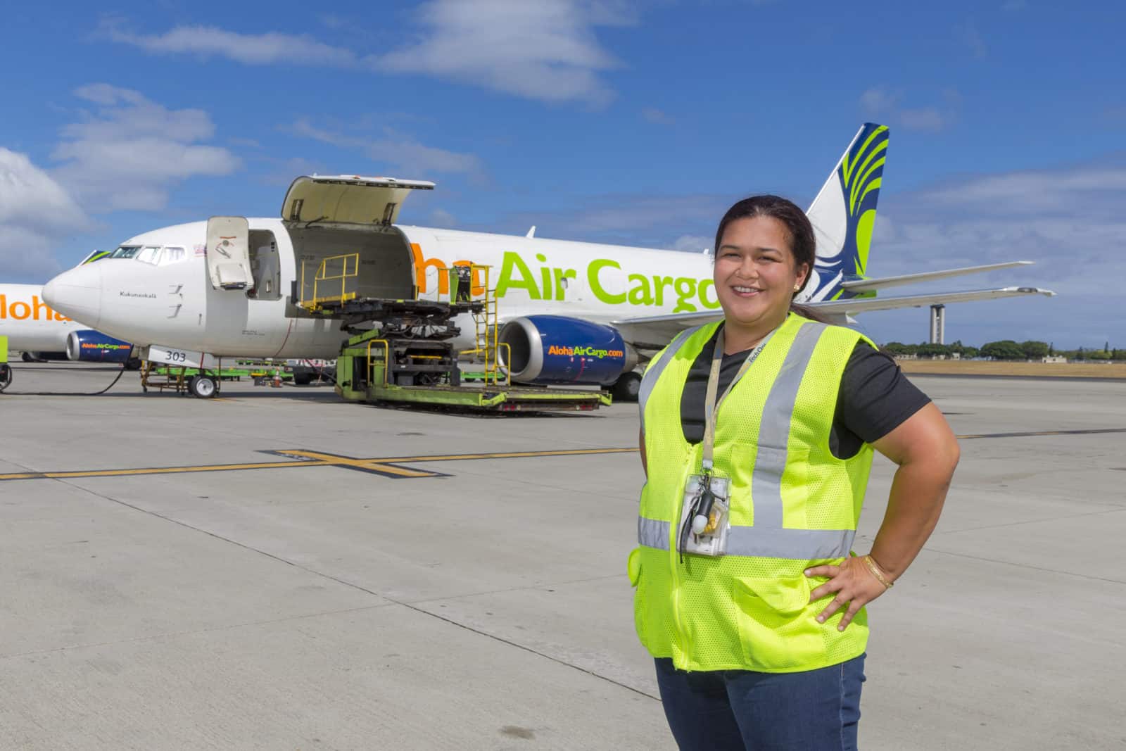 Medeiros stands in a yellow reflective vest, an Aloha Air Cargo plane with bay doors open sits on the tarmac behind her.