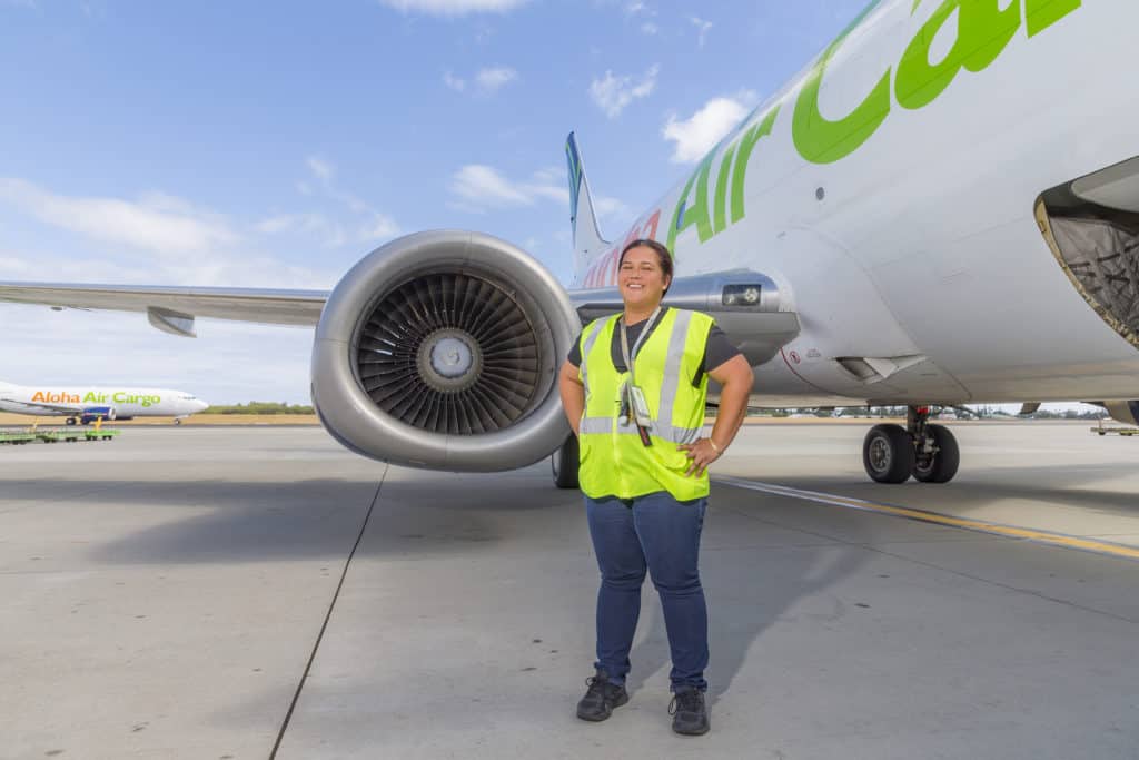 Medeiros has her hands on her hips in front of the engine of an Aloha Air Cargo plane.