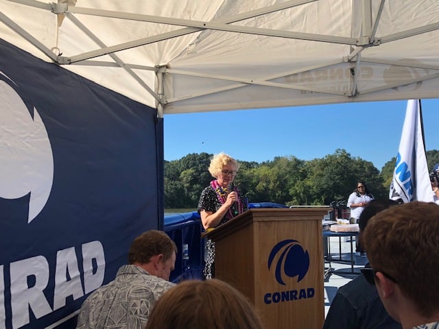 Lynne Parrott stands at a podium with a microphone at Conrad Shipyard.