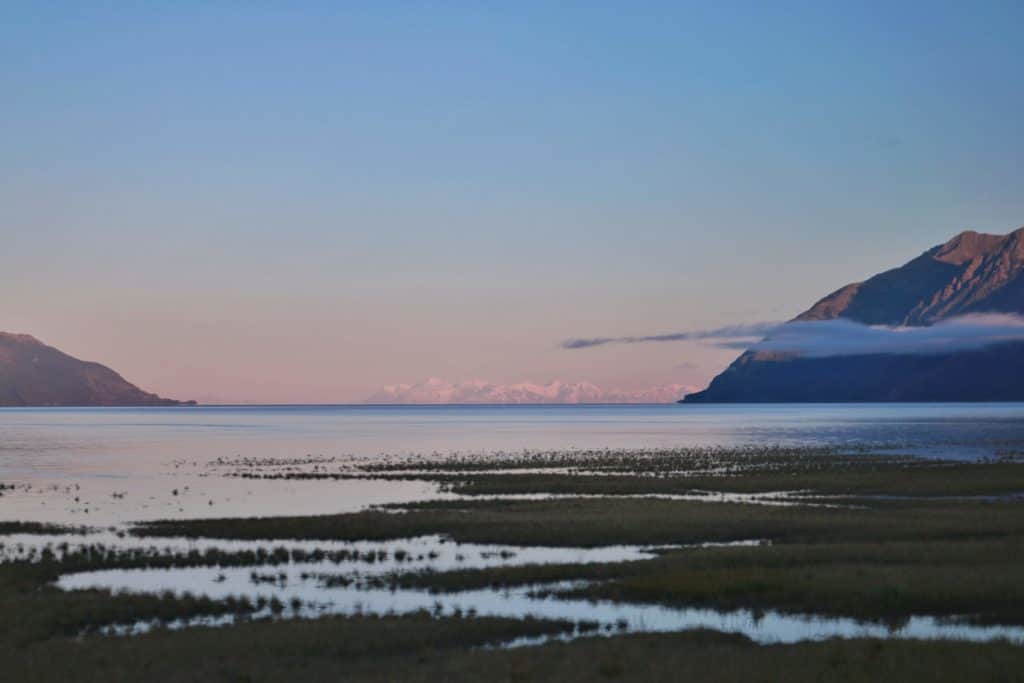 Anchorage landscape shows out of focus marshes leading to a vast expanse of water backed by distant snowcapped mountains. Two hills frame the side of the scene suggesting your gaze to look onward.