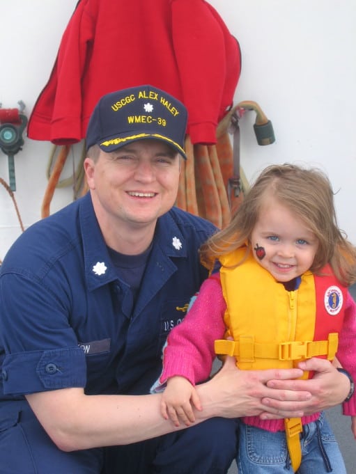 Captain Dixon in a USCG cap holds his young daughter wearing a USCG life jacket.