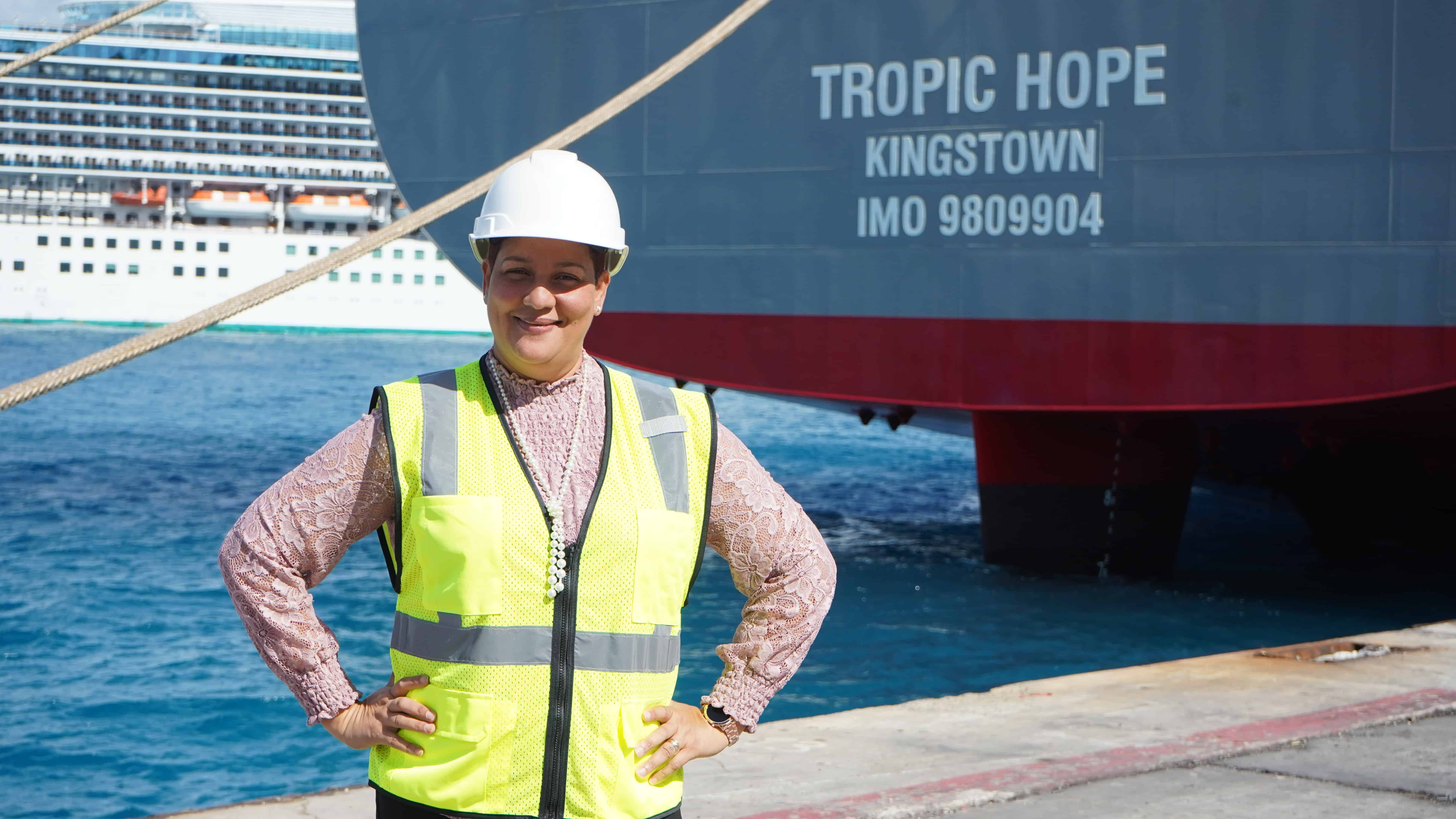 Keyla stands in front of the stern of the Tropic Hope, wearing a hard hat and reflective vest.