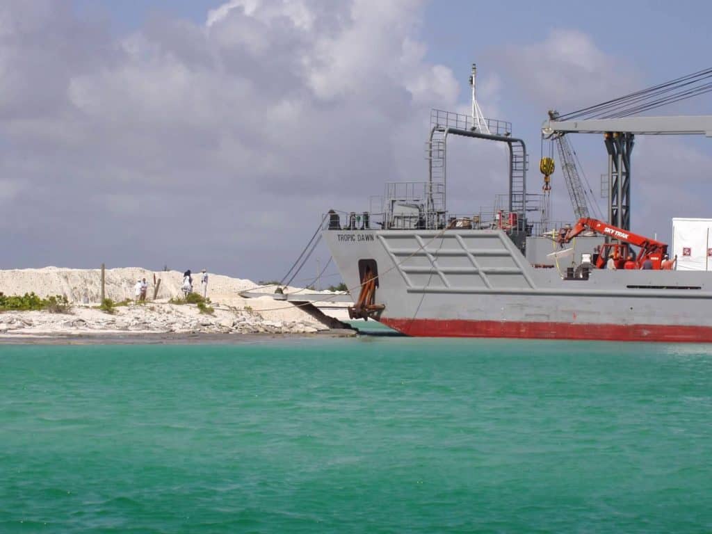 Parrot Cay's loading ramp rests on a sandy beach. The ship is surrounded by shallow blue water.