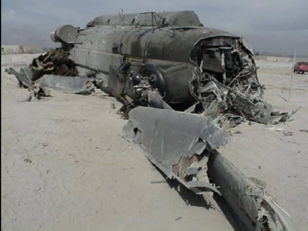 A helicopter, now a hunk of metal and parts lies in a dusty field, almost unrecognizable.