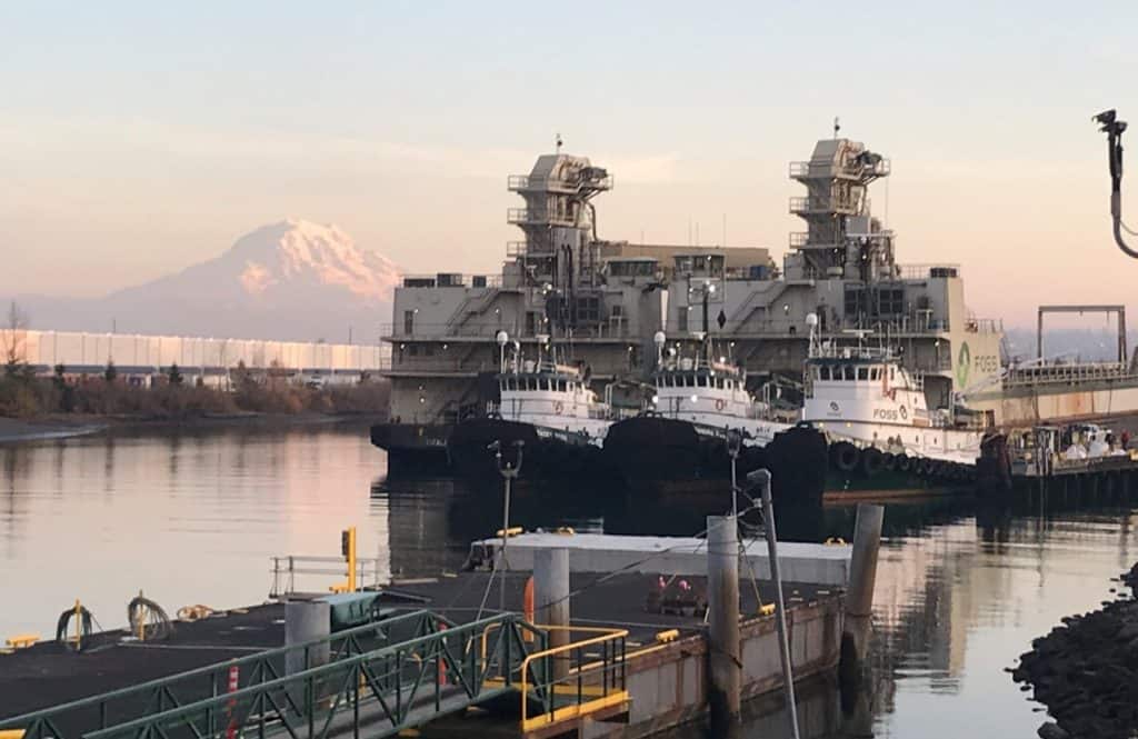 Three Foss tugboats float with their backs to a large Foss barge. A pink Mt. Rainier in the background.