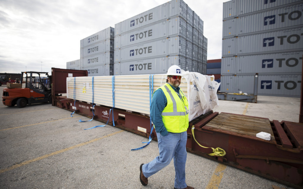 Bennett strolls through TOTE shipyard. Containers stacked high behind him.