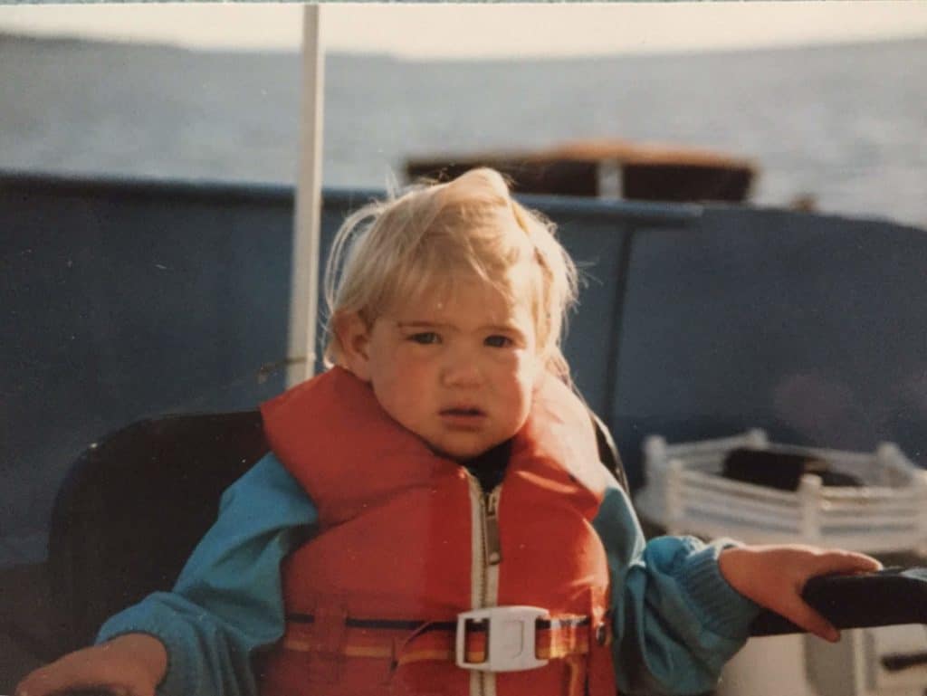 Young Katrina looks at the camera in her oversized life vest holding on to the chair she sits in.