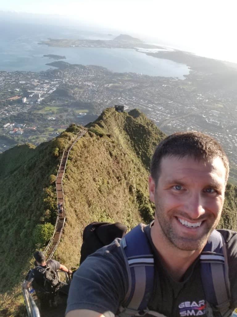 Andy smiles while taking a selfie at the top of a mountain, a city splays out behind him.