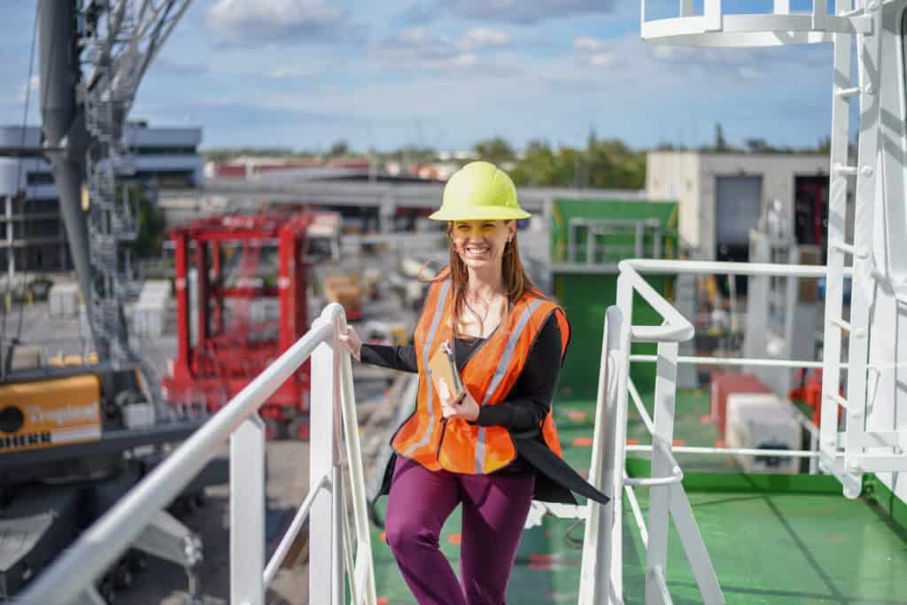Krista smiles as she walks upstairs on a ship, she's wearing a hard hat and orange reflective vest.