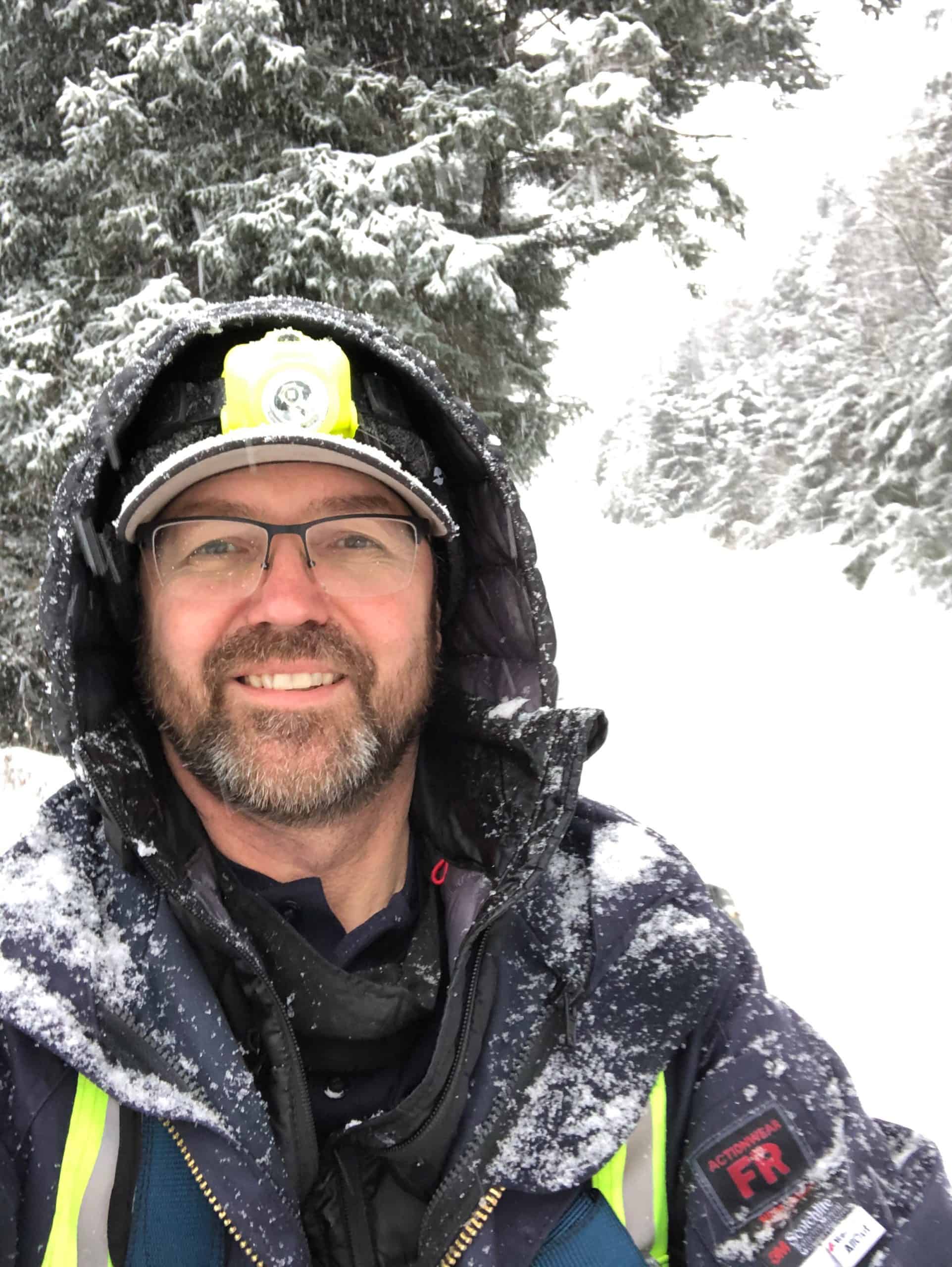 Denker smiles in a heavy jacket and cap, hoodie up, as snow falls around him.