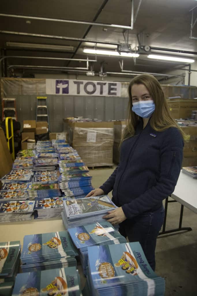 Milena wears a mask in a TOTE warehouse sorting books laid out on the table in front of her.