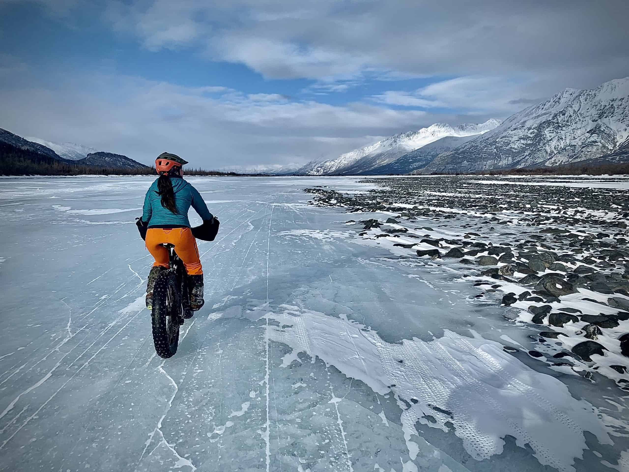 Kelly Willett rides her bike over a frozen lake surrounded by snowy mountains.