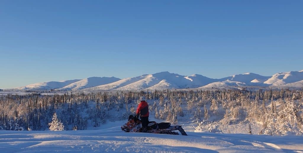 Kurtzweil stands on a snowmobile looking out over a snowy forest and rolling hills of white.