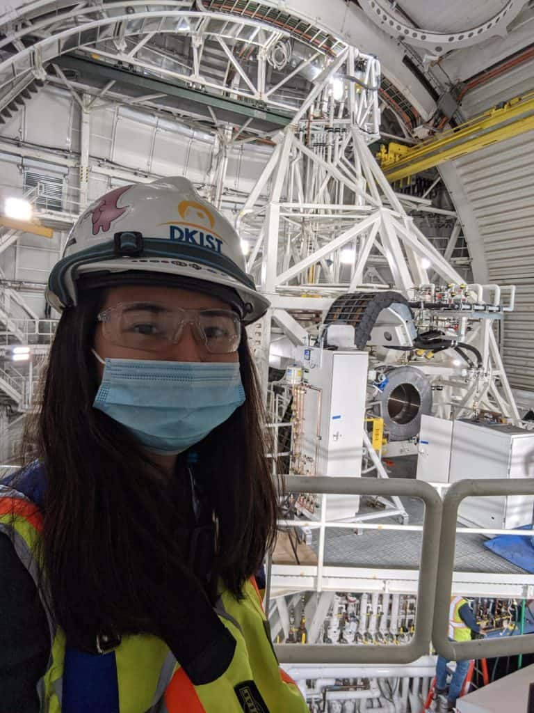 Brialyn wears a mask, hard hat, and yellow reflective vest in a large observatory.