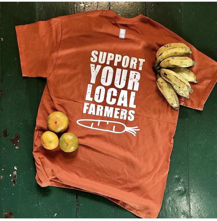 An orange shirt reads, "Support your local farmers." A few bananas and a couple of oranges rest on top of it.