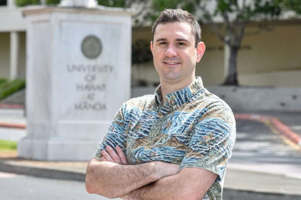 Ben Gershom crosses his arm and smiles in front of a University of Hawaii Statue.