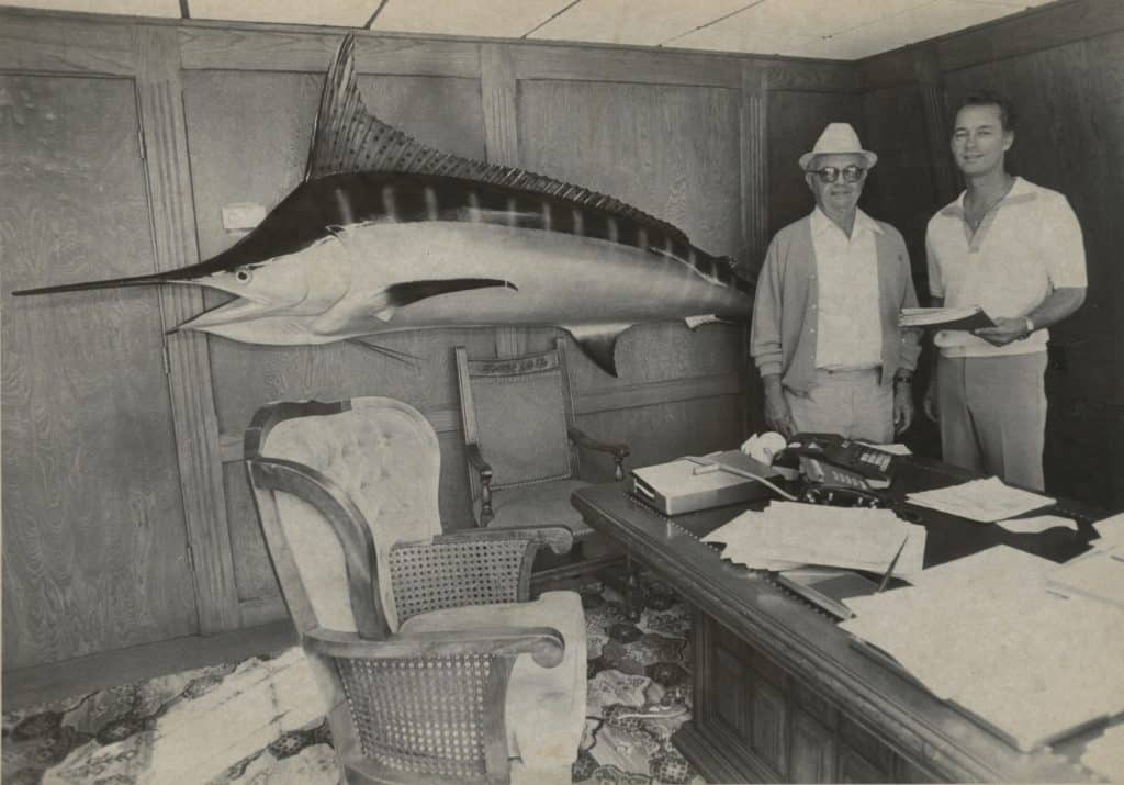 Tom Jankovich Jr. and Sr. pose together in an office with a large marlin on the wall