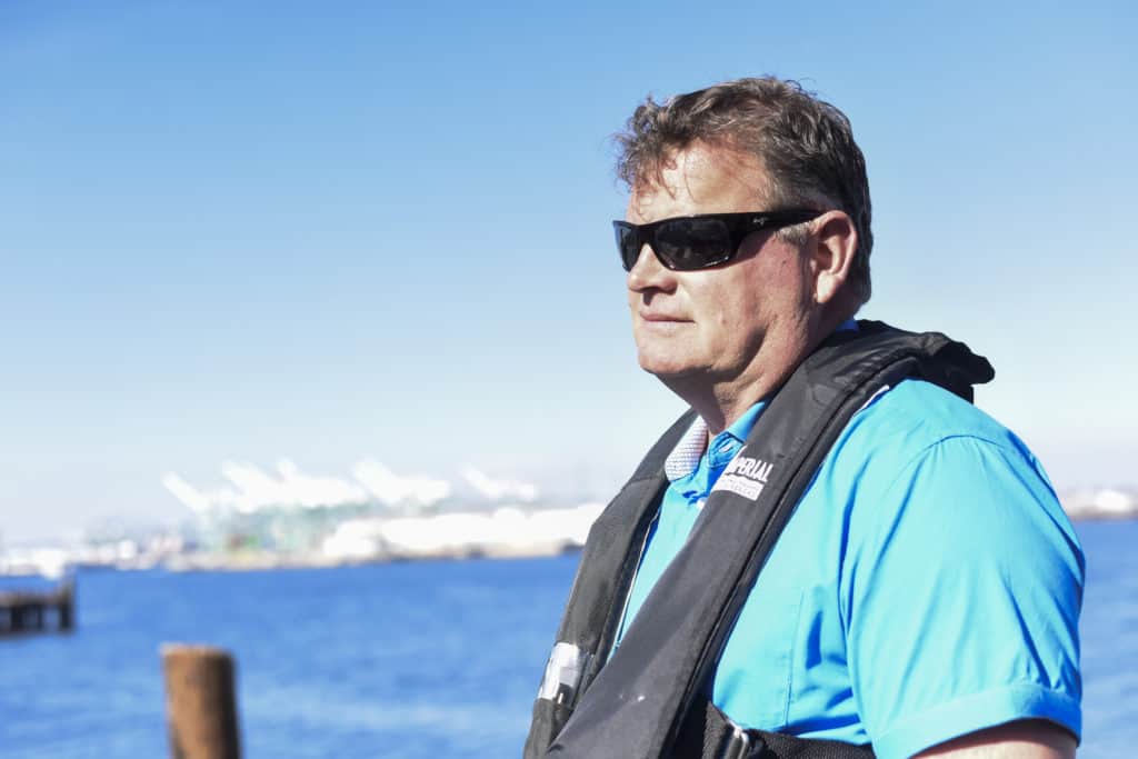 McCaughey wears sunglasses and PFD, a harbor behind him.