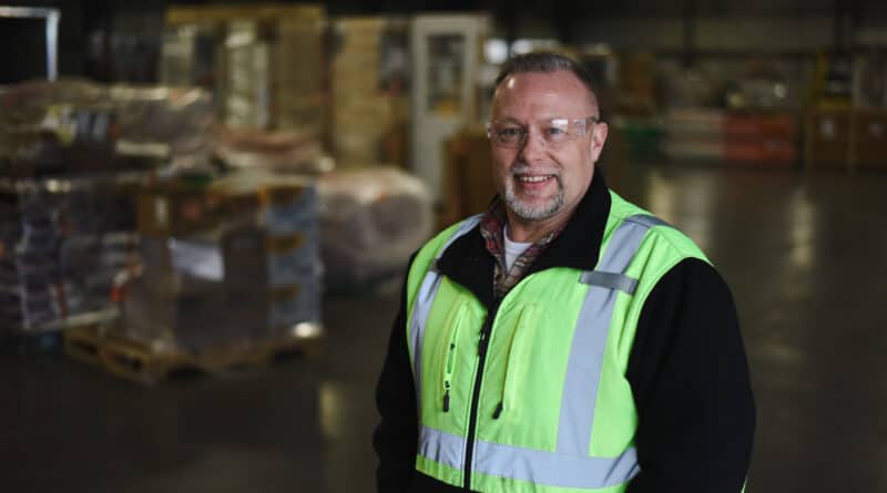 Wendell Hiser smiles in a Carlile warehouse, wearing a green reflective vest and safety glasses.
