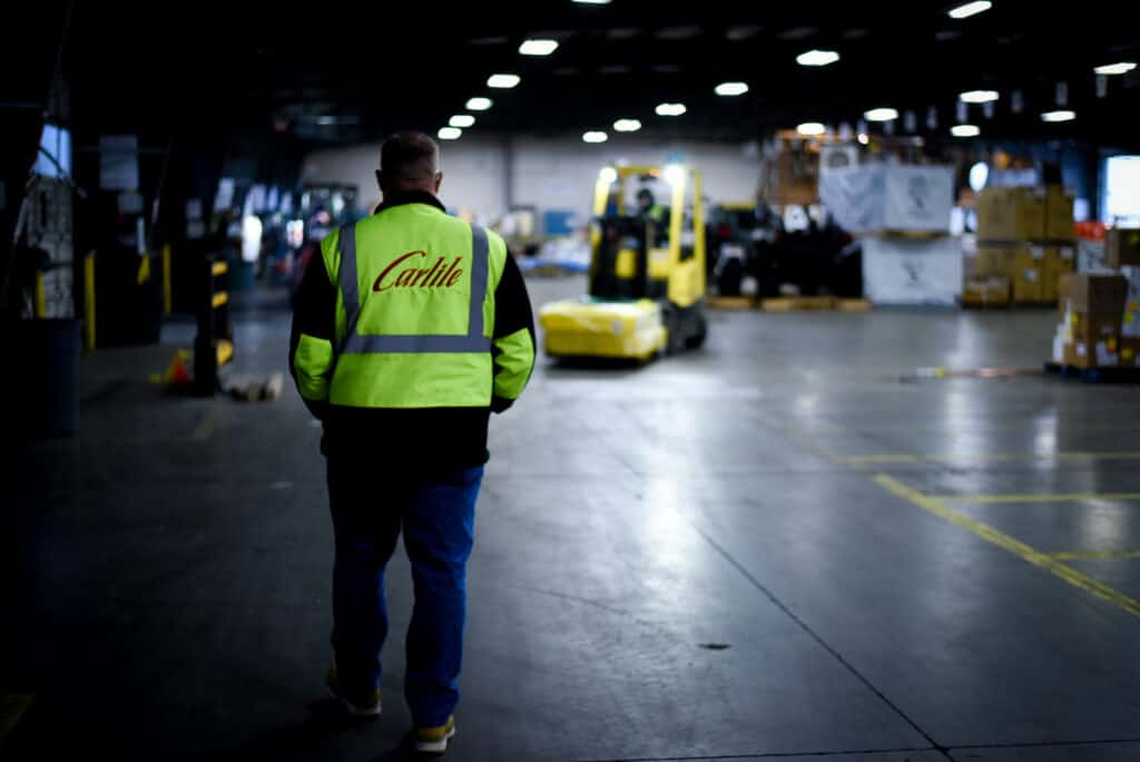 Hiser walks away from the camera in a Carlile reflective jacket. A forklift navigates its way through the Carlile warehouse in the background.