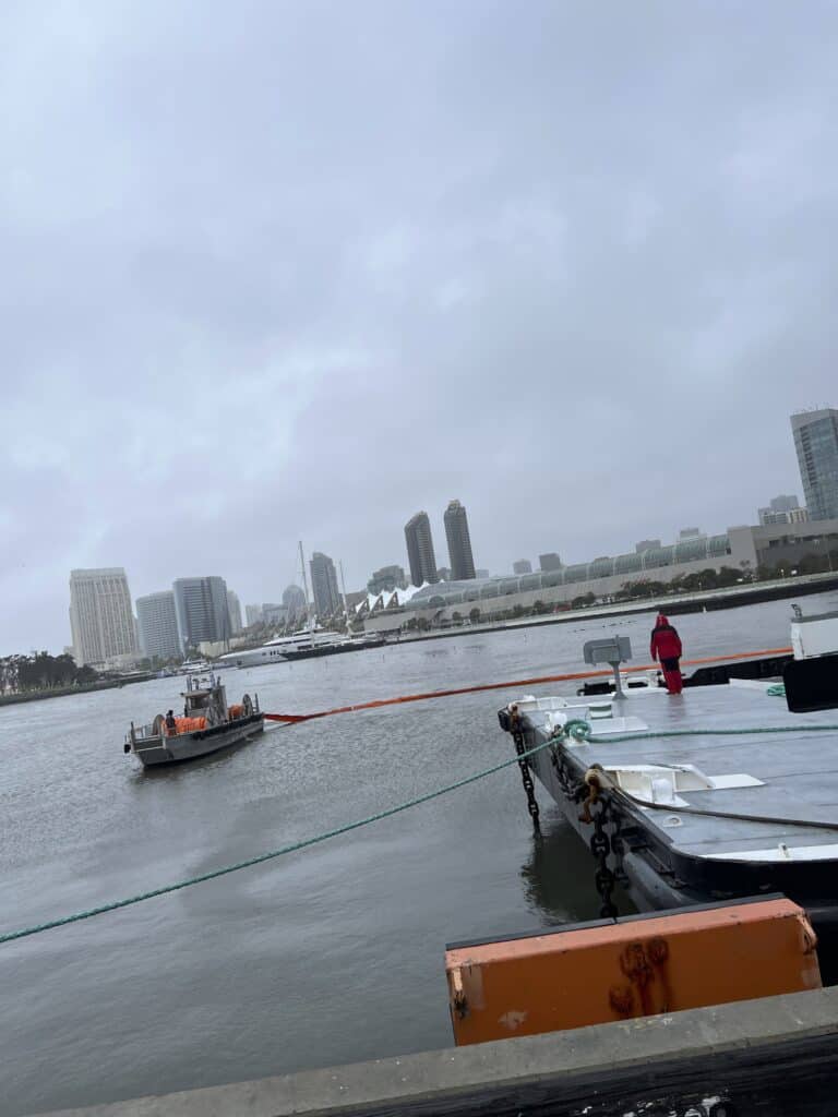 In rainy weather, a small ship circles a barge with a floating line to minimize damage from an oil leak.